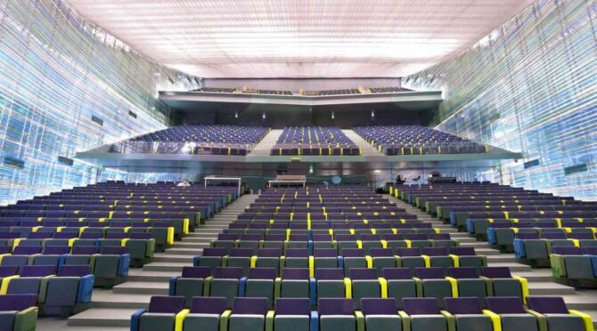 A modern lecture hall with tiered seating, blue chairs, and yellow armrests, framed by translucent, futuristic walls and a diffused light ceiling.