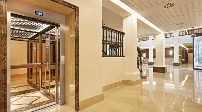 Luxurious elevator lobby with marble walls and ornate ceilings. Three metallic elevators display "1," opening into a grand room with columns and a piano.