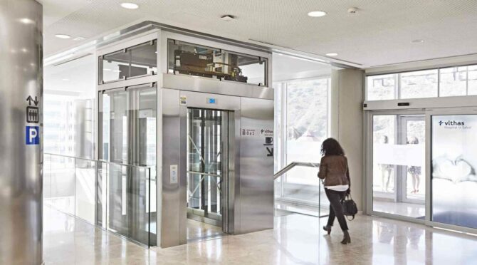 A modern glass and stainless steel elevator in a well-lit hospital lobby, with a woman walking towards the exit.