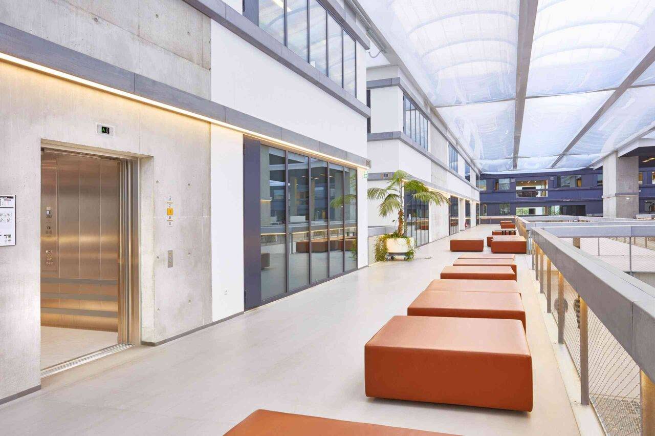 A modern indoor hallway with high ceilings, natural light, orange seating, and glass features.