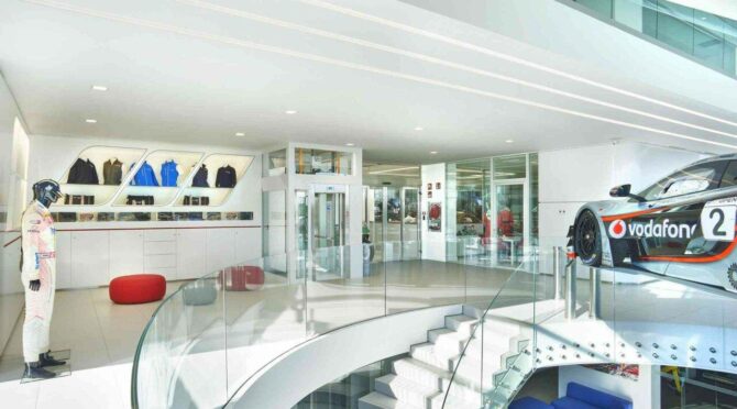 A modern showroom space with glass railings on the staircase, a mannequin in racing gear, clothing displays, a red ottoman, a glass door, and a race car with Vodafone branding.