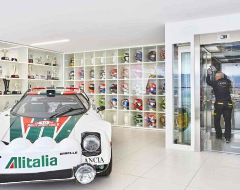 A room displaying trophies and racing helmets on shelves, with a white rally car in the foreground featuring "Alitalia" and "Rallye Monte Carlo" text. A person works on an elevator to the right.