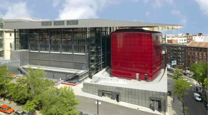 A modern building with a bold red structure, glass and metal facade, surrounded by trees and parked cars.