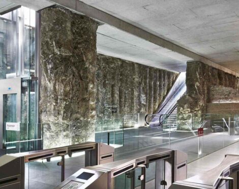 A modern underground station with exposed rock walls, a glass elevator, turnstiles, and an escalator.